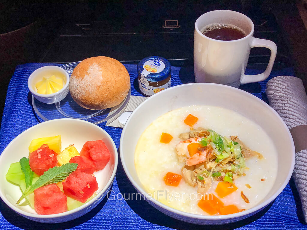 A dinner tray with a blue tablecloth carries a bowl of congee accompanied by a bowl of cut fruits and a roll with butter and jam.