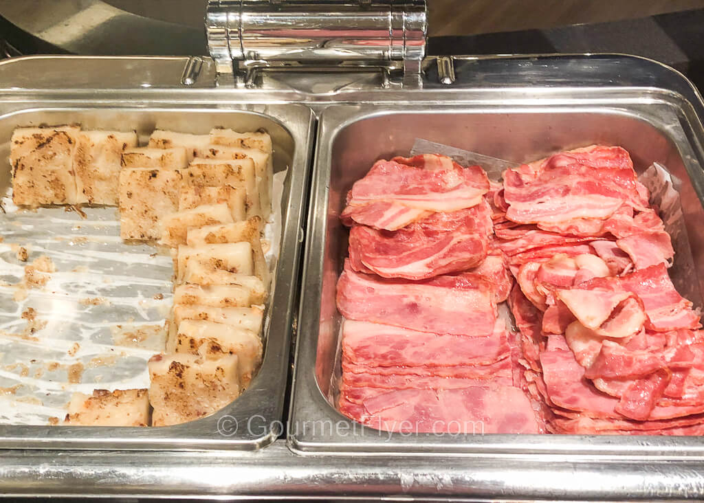 A pair of chaffing dishes are set up on a counter with turnip cakes to the left and bacon to the right.