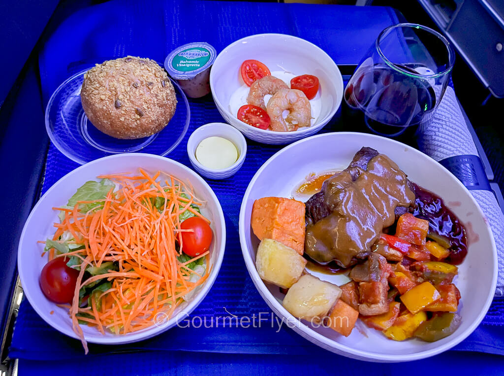 An airplane first class dinner tray contains a beef entree, side salad, a shrimp appetizer and a glass of red wine.