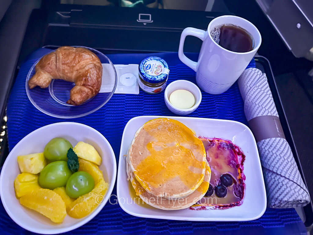 A dinner tray contains a plate of pancakes, a bowl of cut fresh fruits, a croissant and a cup of black coffee.