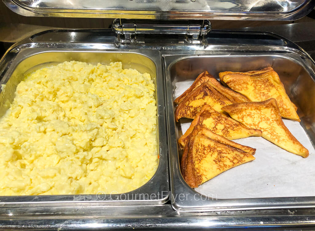 A chaffing dish filed with scrambled eggs is accompanied by a few slices of French toast to its right.