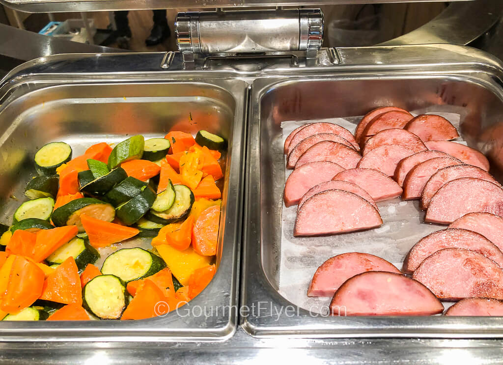 Two chaffing dishes are placed side by side, one with mixed sauteed vegetable and the other with grilled ham.