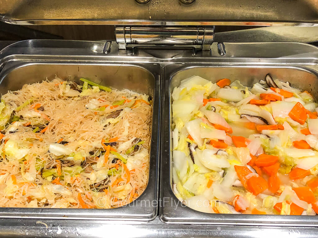 A pair of chaffing dishes contain vermicelli on the left and mixed vegetables with cabbage and carrots on the right. The.
