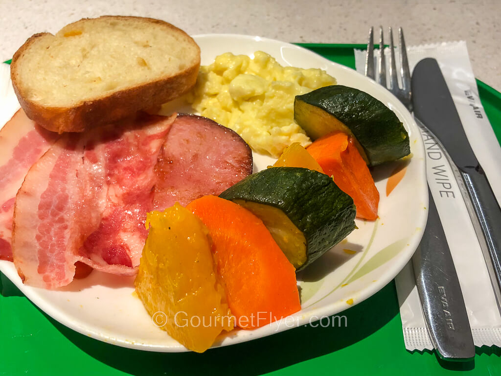 A breakfast plate that contains eggs, ham, vegetables and bread is placed on a tray with silverware.