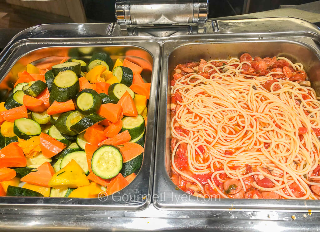 The chaffing dish on the left contains zucchinis, squash, and carrots while the one on the right contains spaghetti in a red sauce with sliced sausages. 