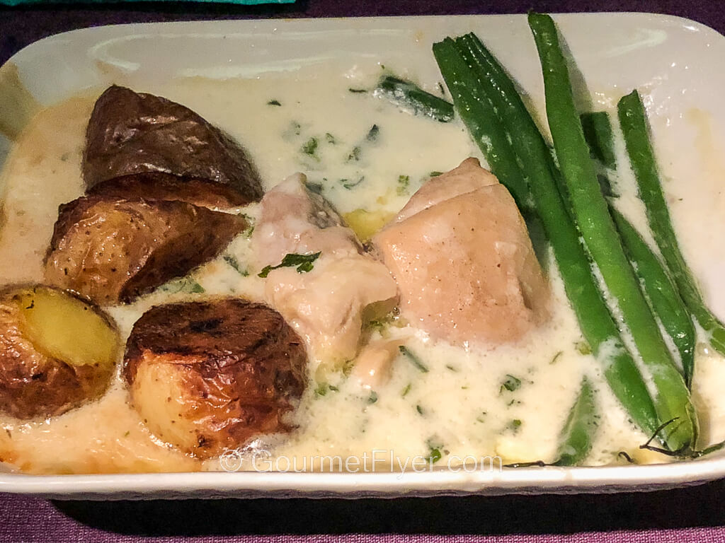 An airline dinner dish contains chunks of white chicken meat braised in a white sauce and accompanied by long string beans and roasted potatoes.
