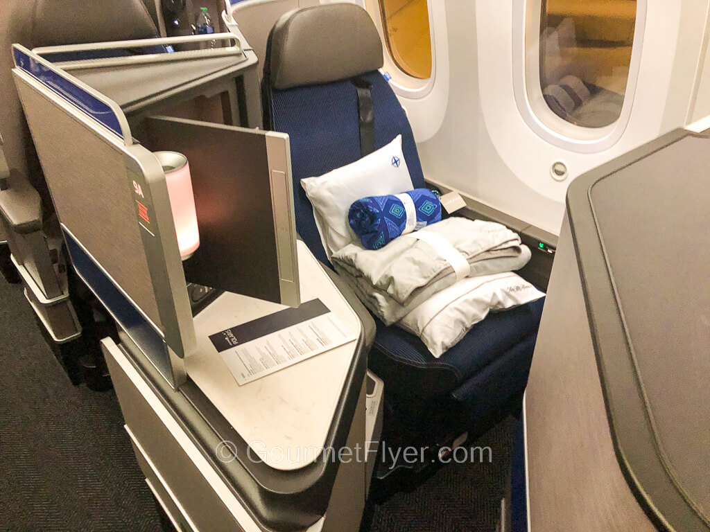 A business class seat with two windows to its left has a stack of blankets and beddings placed on it.