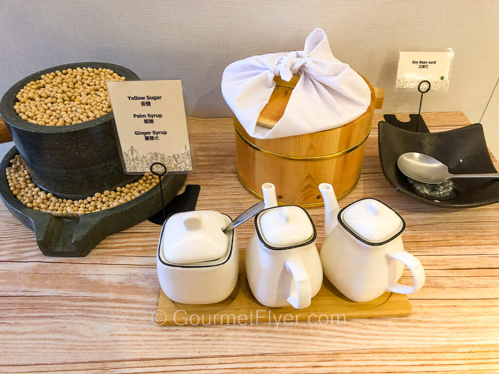 A station is set up with soybean curds in a wooden bucket, syrups in mini teapots, and sugar in a ceramic condiment container.