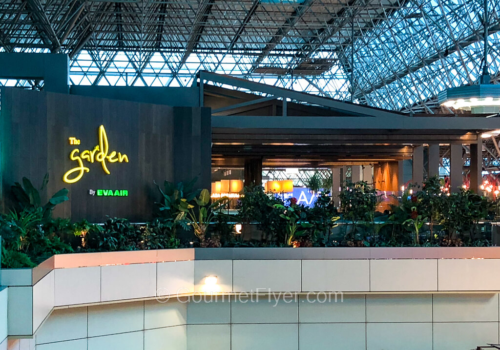 A trendy looking space under an airport terminal's glass ceiling has yellow lighted signs identifying it as The Garden.