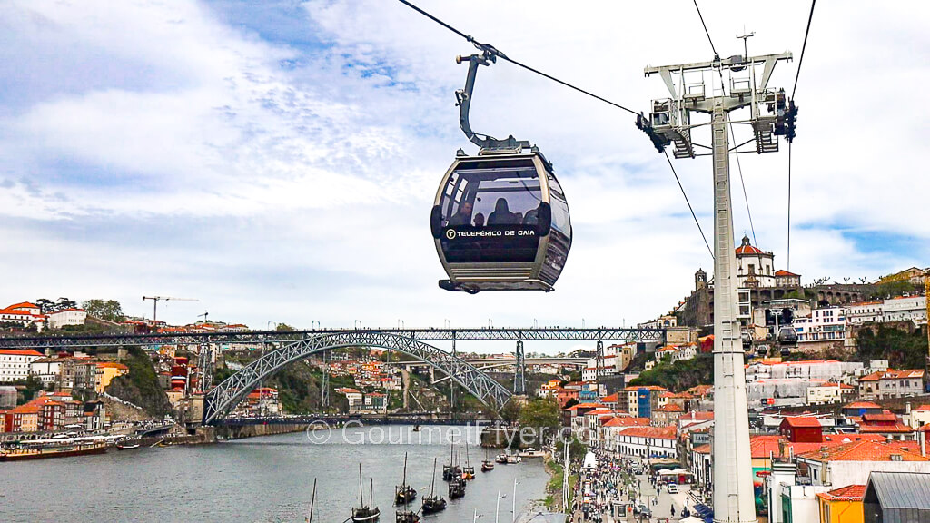 A cable car is traveling above the streets of the Gaia Riverside, with the Luis I Bridge in the background.