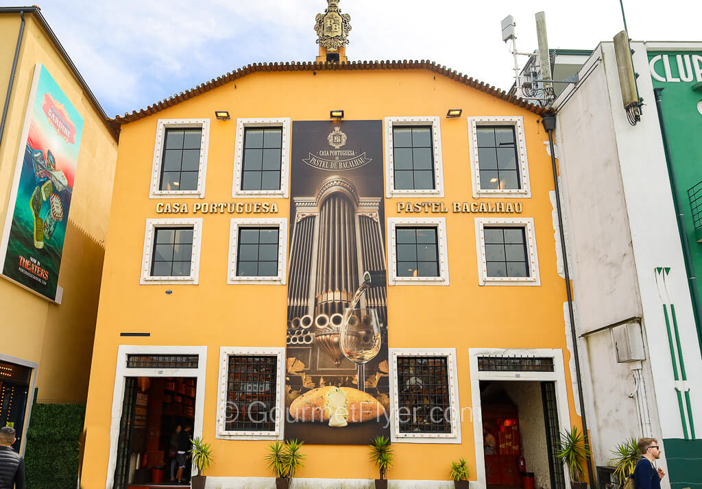 A three-story traditional building has a mustard yellow facade with a painting of a pastry.