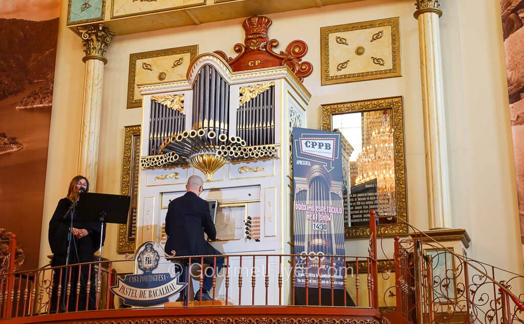 A man plays a large while pipe organ while accompanied by a female singer standing alongside him.