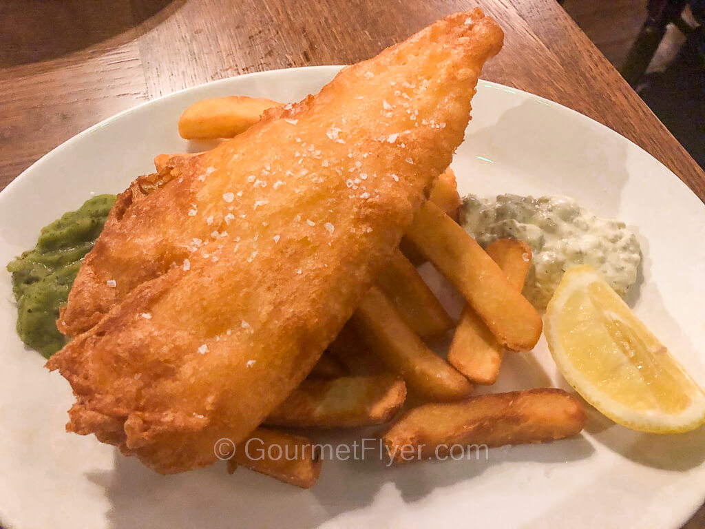 A large piece of fried fish filet sits atop a stack of French fries accompanied by a lemon wedge.