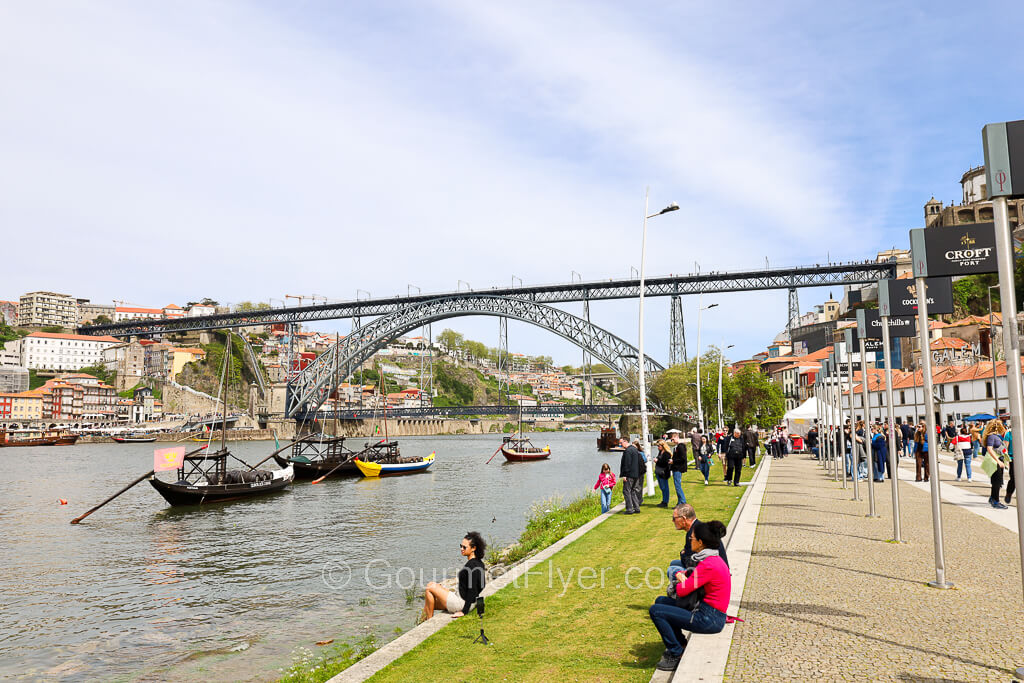 Tourists walk along the grassy waterfront of Gaia is on the right side of the river, across the bridge in the background.