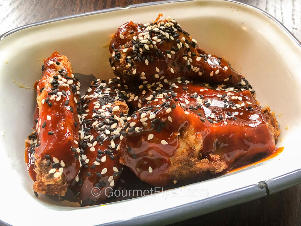 A small serving bowl contains chicken wings drenched in a thick reddish color sauce and sprinkled with sesame.