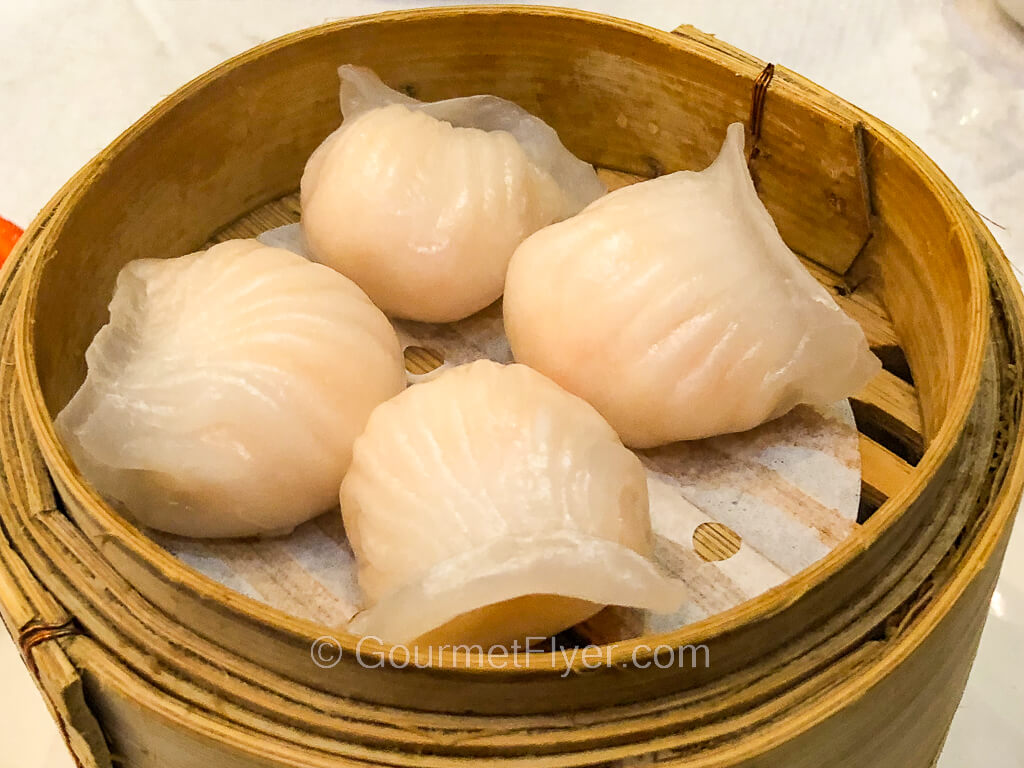 Four shrimp dumplings in white wrappers are served in a bamboo steamer.