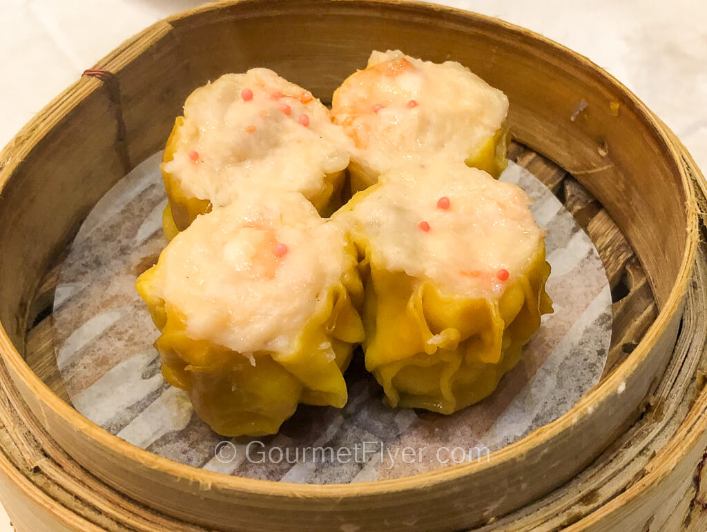 Four dumplings in yellow wrappers all stuck together two by two are served in a bamboo steamer.
