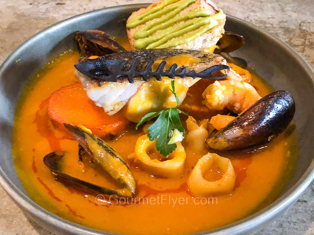 A stew of seafood like fish filet, squid, mussels, etc. are served in an orange color broth.