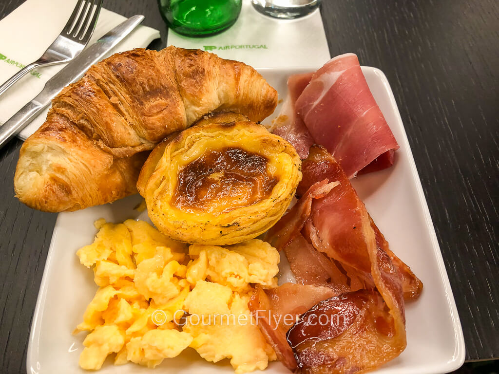 A small plate contains scrambled eggs accompanied by various meats and an egg tart in the center.