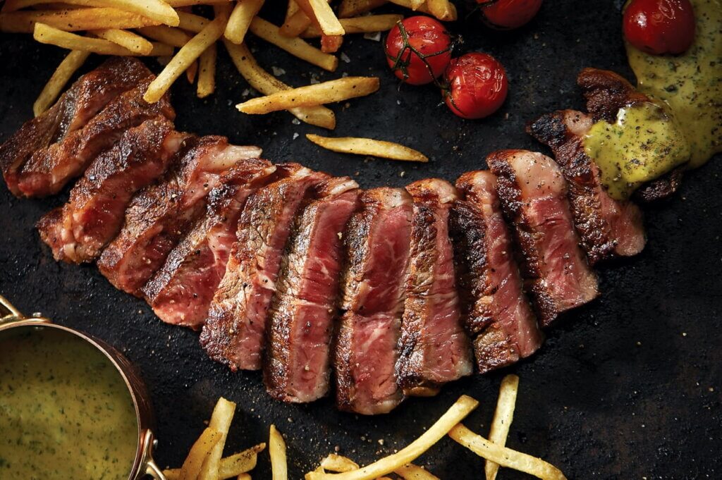 Sliced medium rare steaks are served on a platter sprinkled with French fries and garnished with cherry tomatoes.