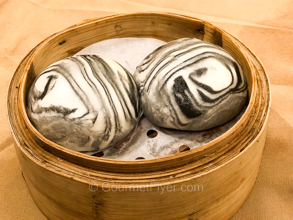 A dim sum steamer contains two buns with a black and white marbled dough.