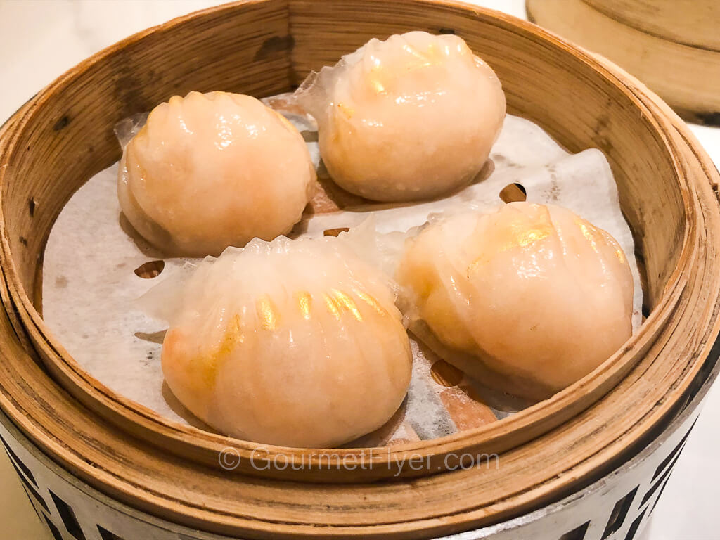 A bamboo steamer contains four dumplings in white skins with a brush of gold.