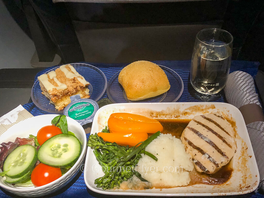 Review of United's First Class flight features a dinner set with chicken, side salad, cake for dessert, and a glass of red wine.