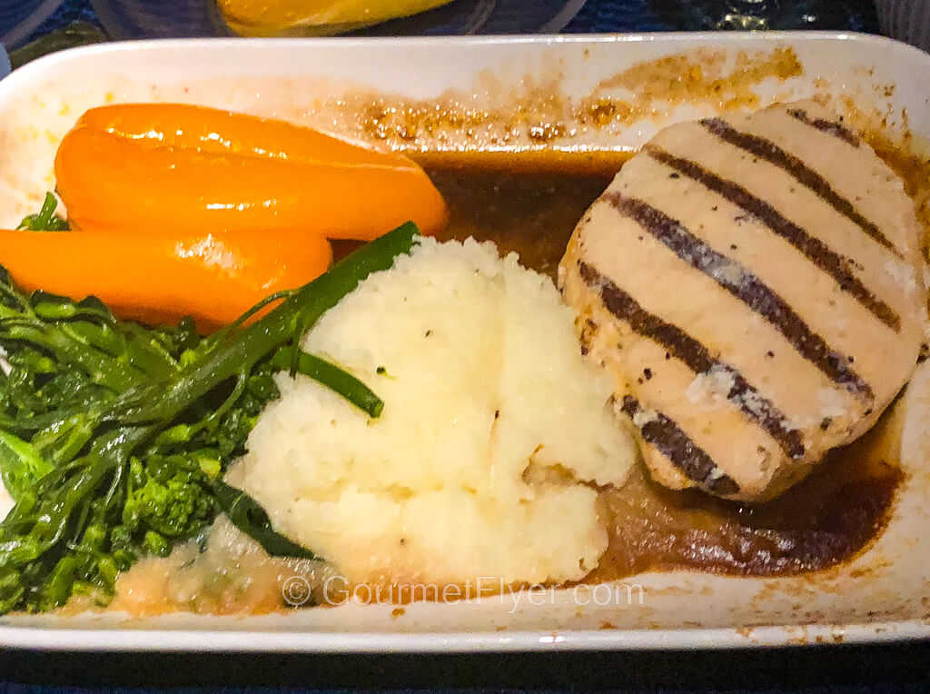 A dinner dish contains a grilled chicken filet served with brown gravy, mashed potatoes, broccolini, and orange bell peppers.