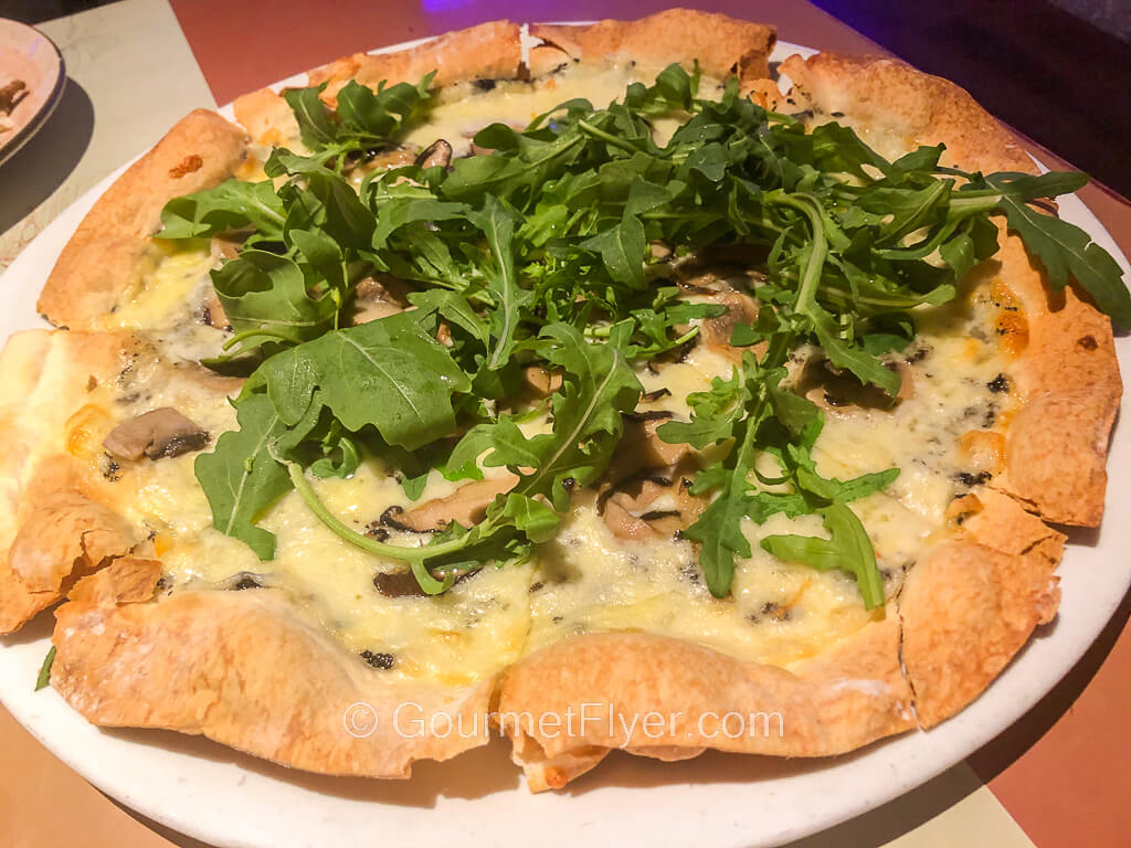 A thin pizza is topped with a pile of arugula leaves and is served on a round plate.