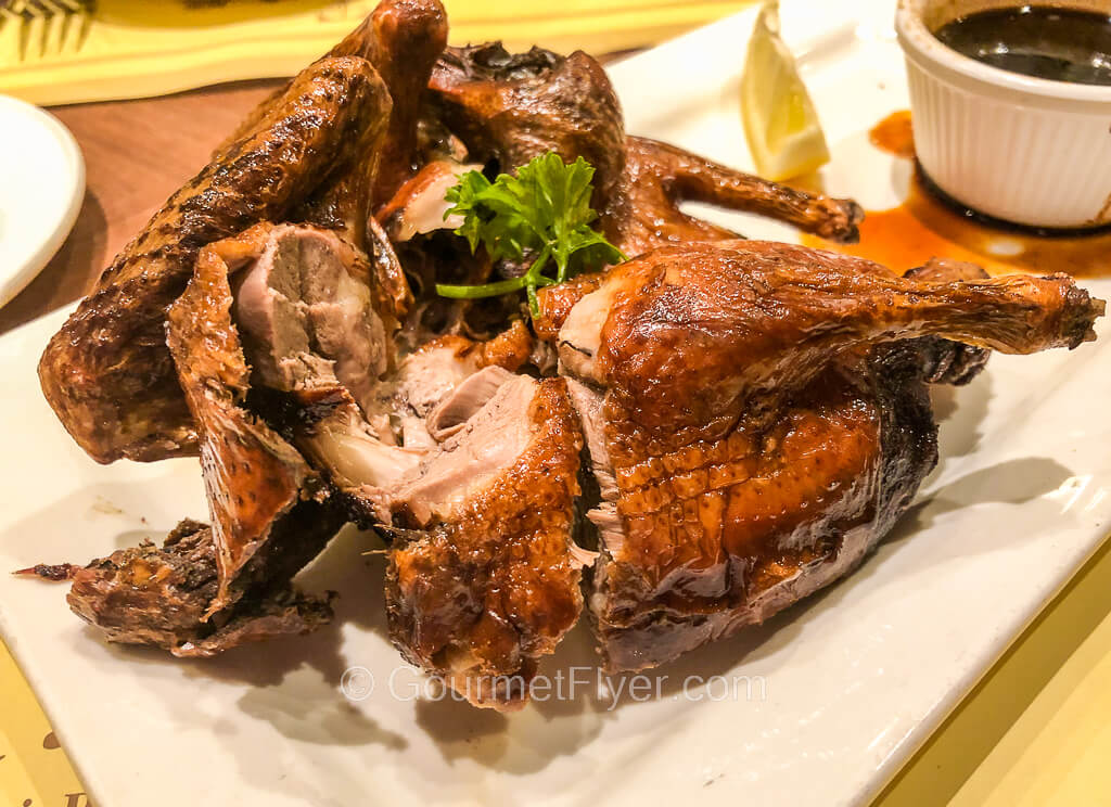 A roasted squab is cut into four quarter pieces and accompanied with a side of dark sauce.