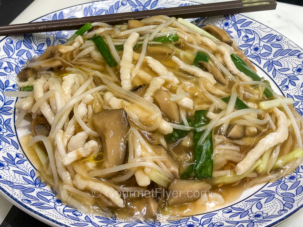 A plate of fried noodles is topped with pork, mushrooms, and plentiful of gravy.