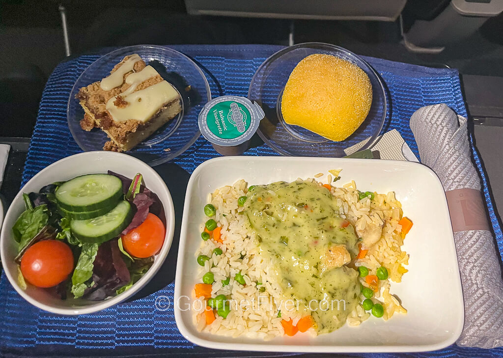 An airline dinner tray contains a chicken dish with rice and green gravy, a side salad, roll, and a piece of cake.