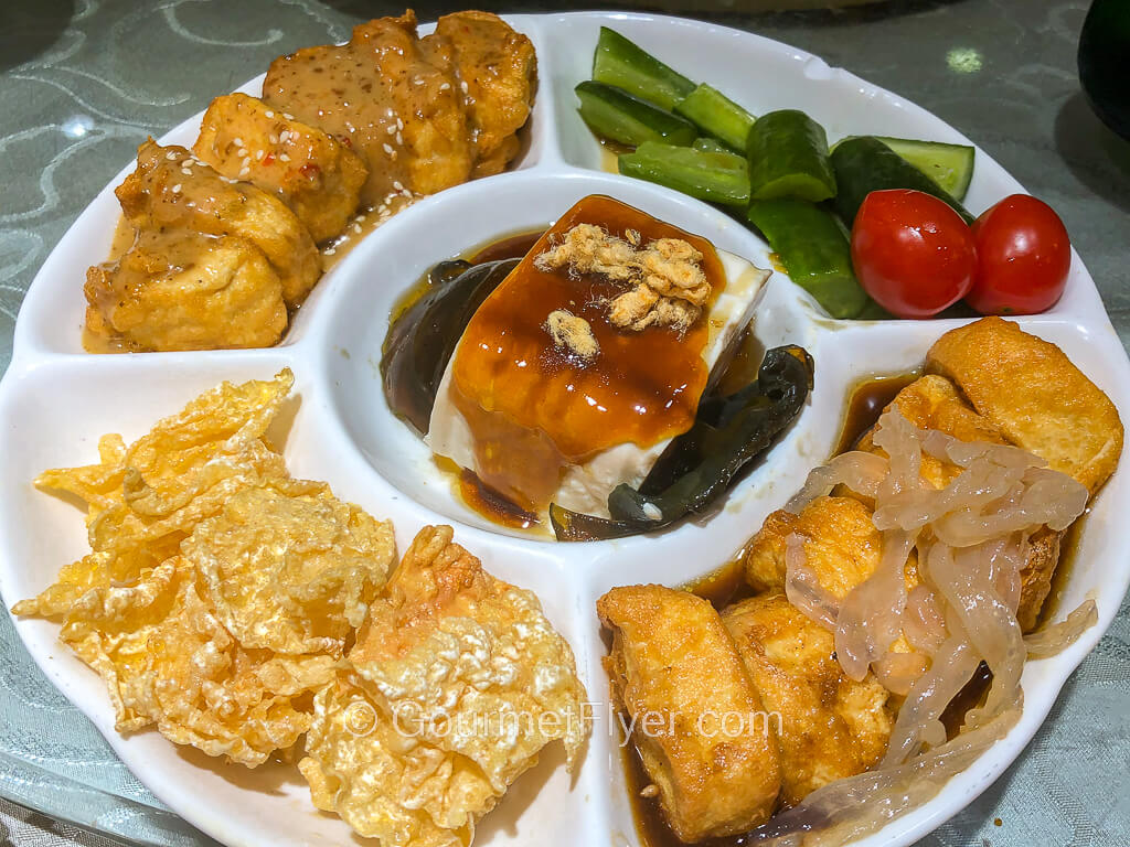 A partitioned circular platter contains various forms of bean curd and are garnished with cherry tomatoes and cucumbers.