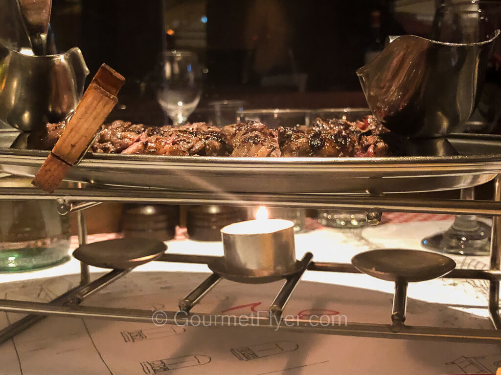 A metal platter containing pieces of meat is placed on a rack over a chafing candle.