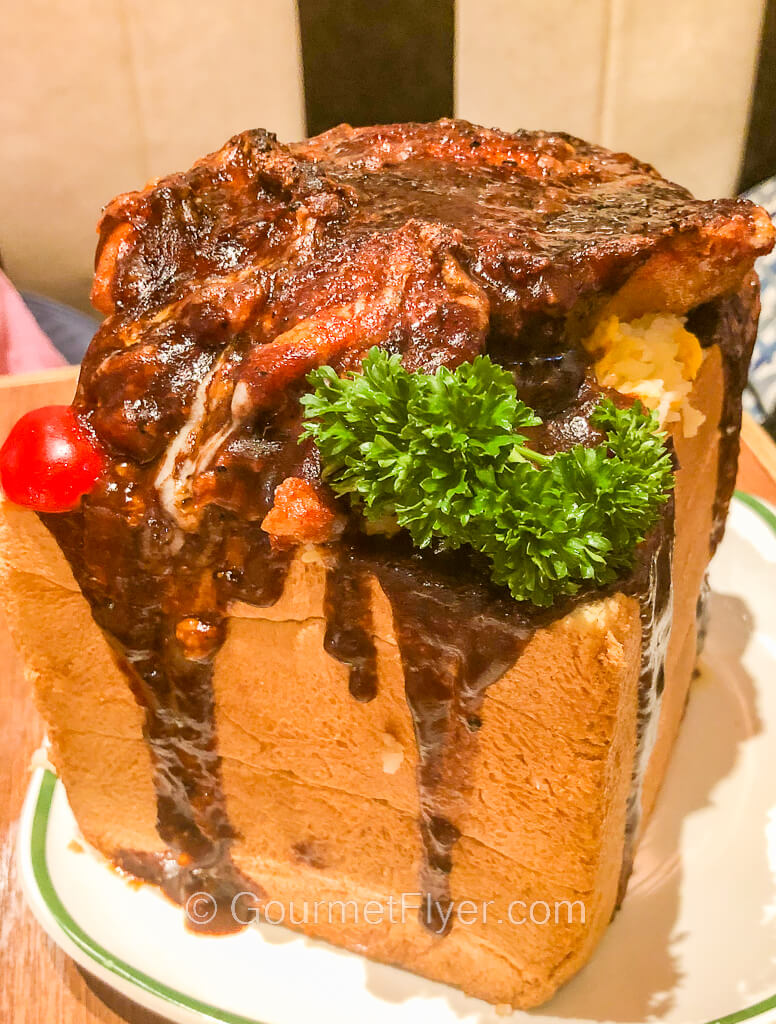 A slice of pork chop covered in black pepper sauce is placed on top of a loaf of bread that looks like a cubic brick.