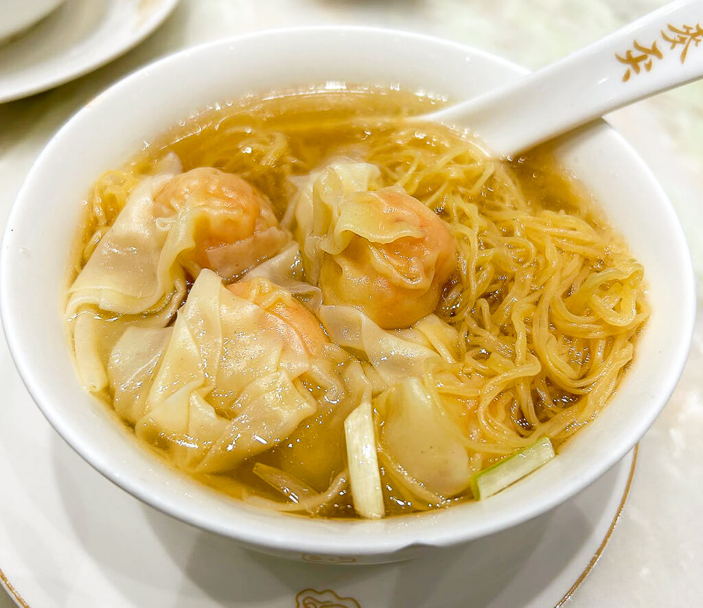 Four wonton dumplings sit atop a bowl of noodle soup with egg noodles in a yellowish broth.