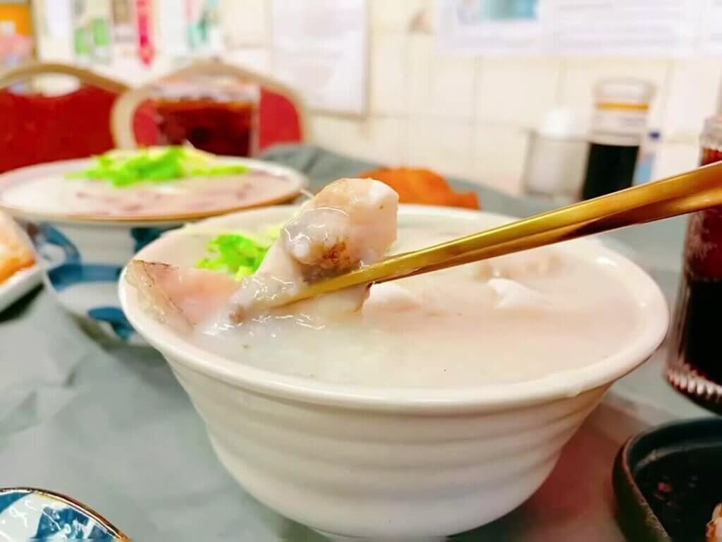 A pair of chopsticks picks up a piece of fish filet from a bowl of congee.