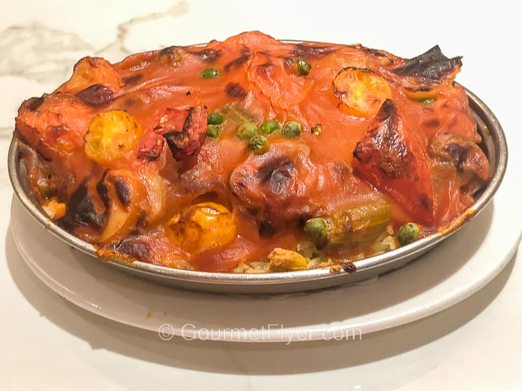 An oval metal baking dish is filled with a cheesy red sauce topped with tomatoes and other vegetables.