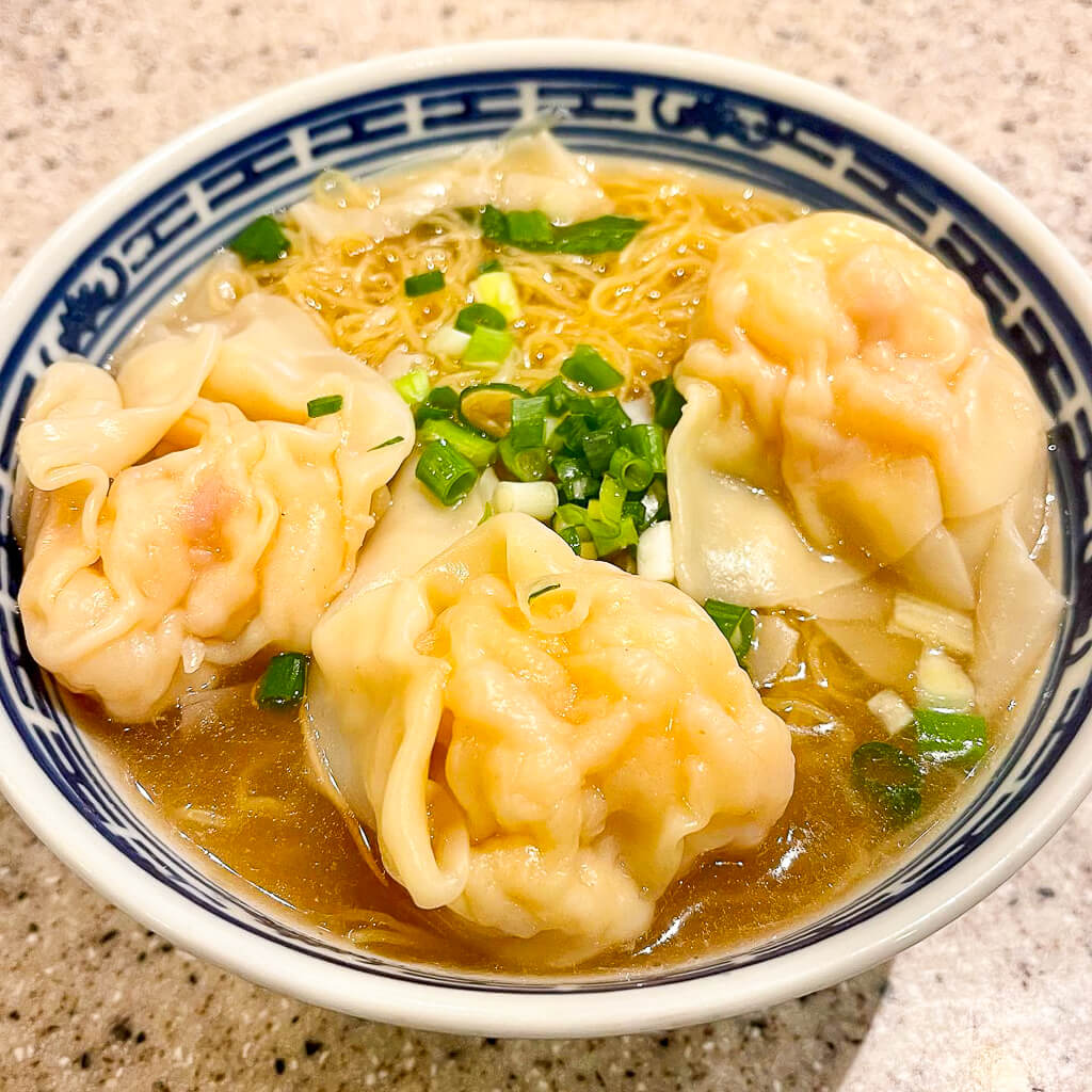 Three large wonton dumplings sit on top of a bowl of noodle soup sprinkled with green onions.
