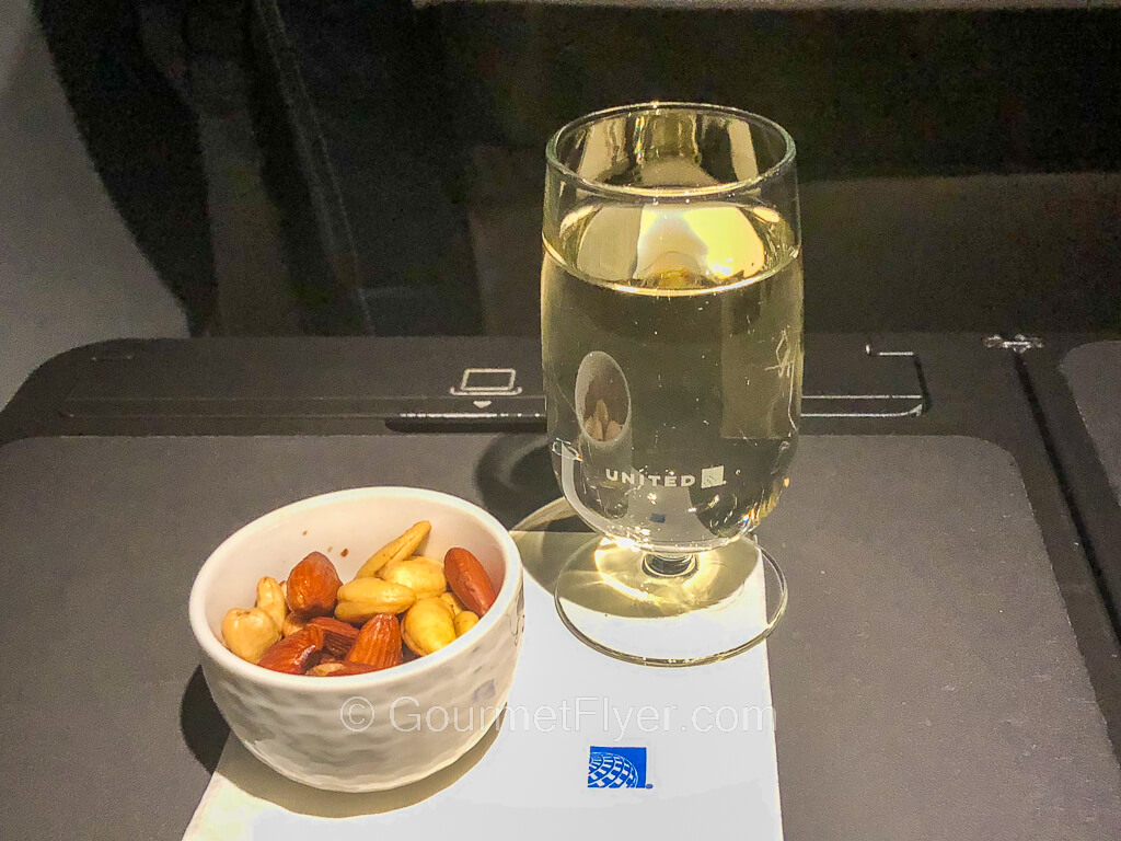 A glass of white wine is served in a glass with a "United" logo and is accompanied by a ramekin filled with mixed nuts.