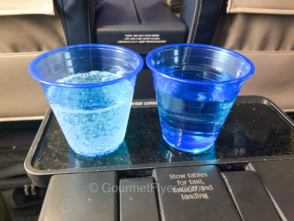 Two blue plastic cups are placed side by side on an airline seat table, one contains water and the other contains sparkling wine.