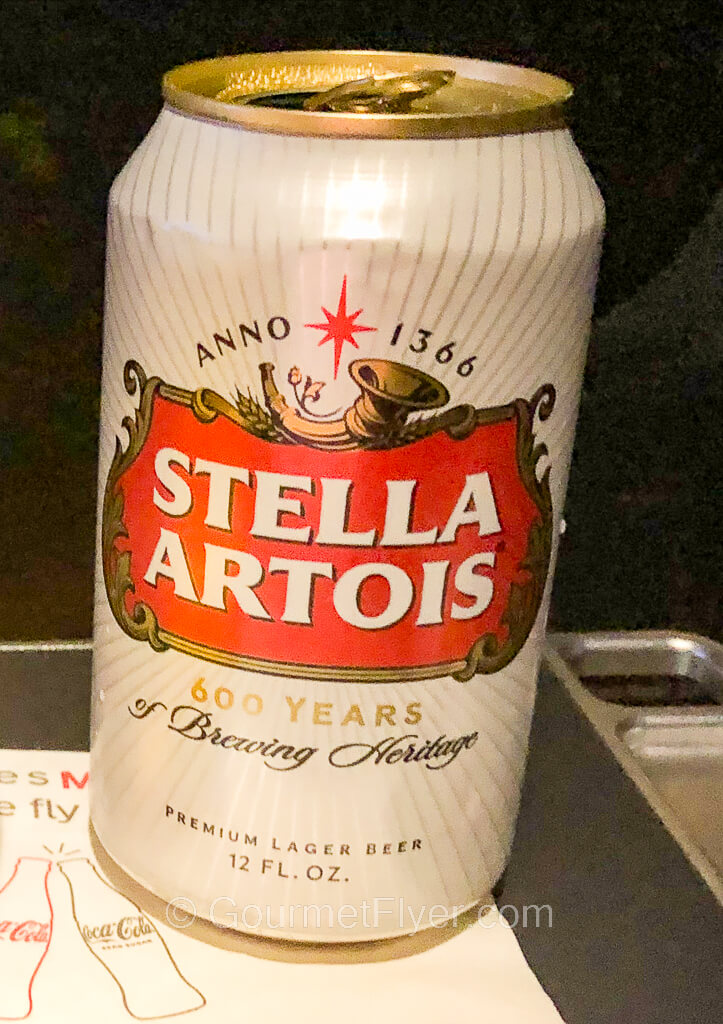 A can of opened Stella Artois beer sits on a napkin atop a tray table.