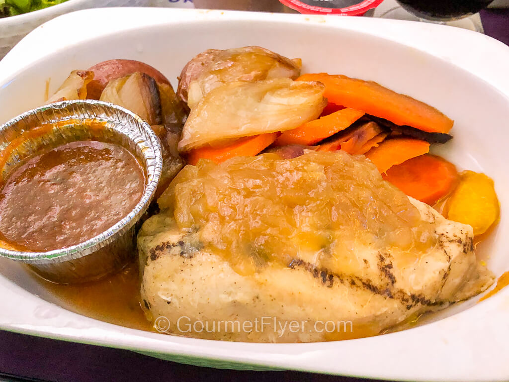 A grilled piece of meat is served with carrots, potatoes, and a side of a brown sauce in a tin cup.