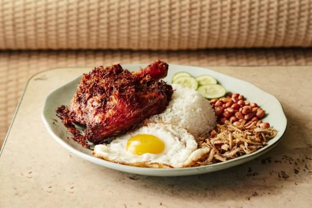 A large piece of meat with a burnt skin is served with white rice, a fried egg, and other garnishments.