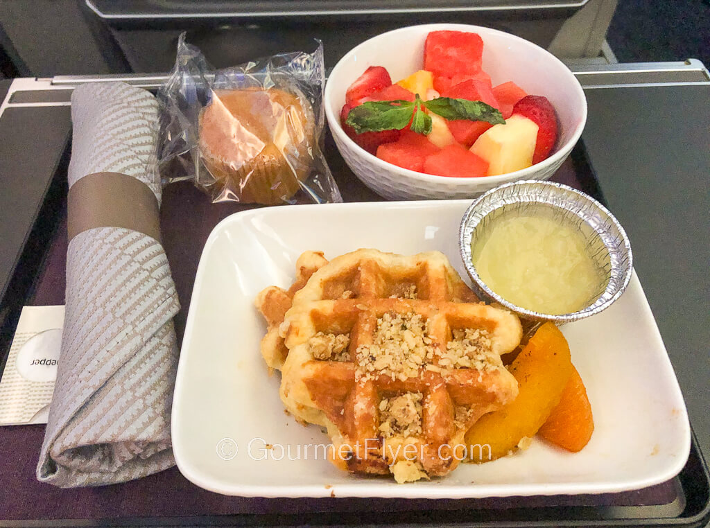 Waffles with slices of fruits and sauce is served with a bowl of cut fresh fruits and a muffin.