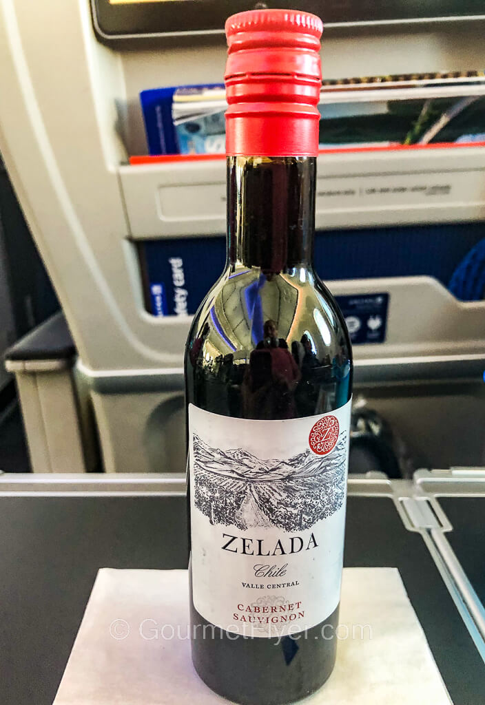 A mini bottle of red wine is placed on a tray table with a seatback in the background.