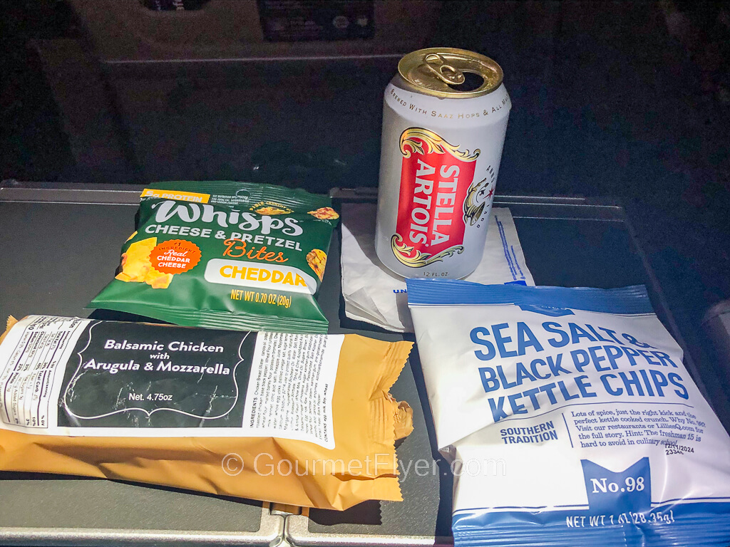 Spread out on a tray table are a wrapped sandwich, packets of chips, and a can of Stella beer.