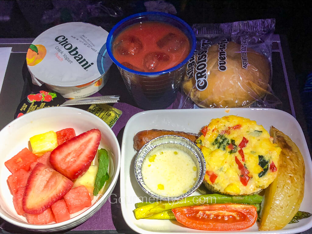 A breakfast tray contains a colorful egg dish garnished with asparagus spears, a bowl of fruits which include strawberries, and a pastry wrapped in plastic.