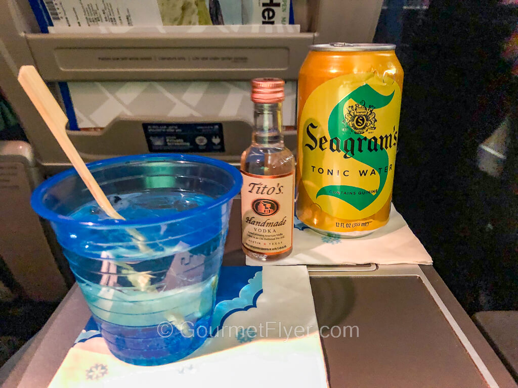 A drink in a blue plastic cup is accompanied by a mini bottle of liquor and a can of tonic water.