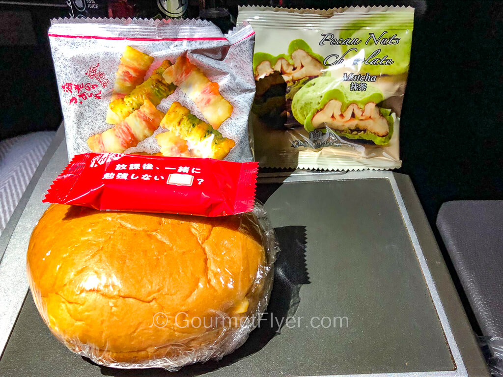 A sandwich made with a bun is accompanied by a kit kat and packets of snacks on a tray table.